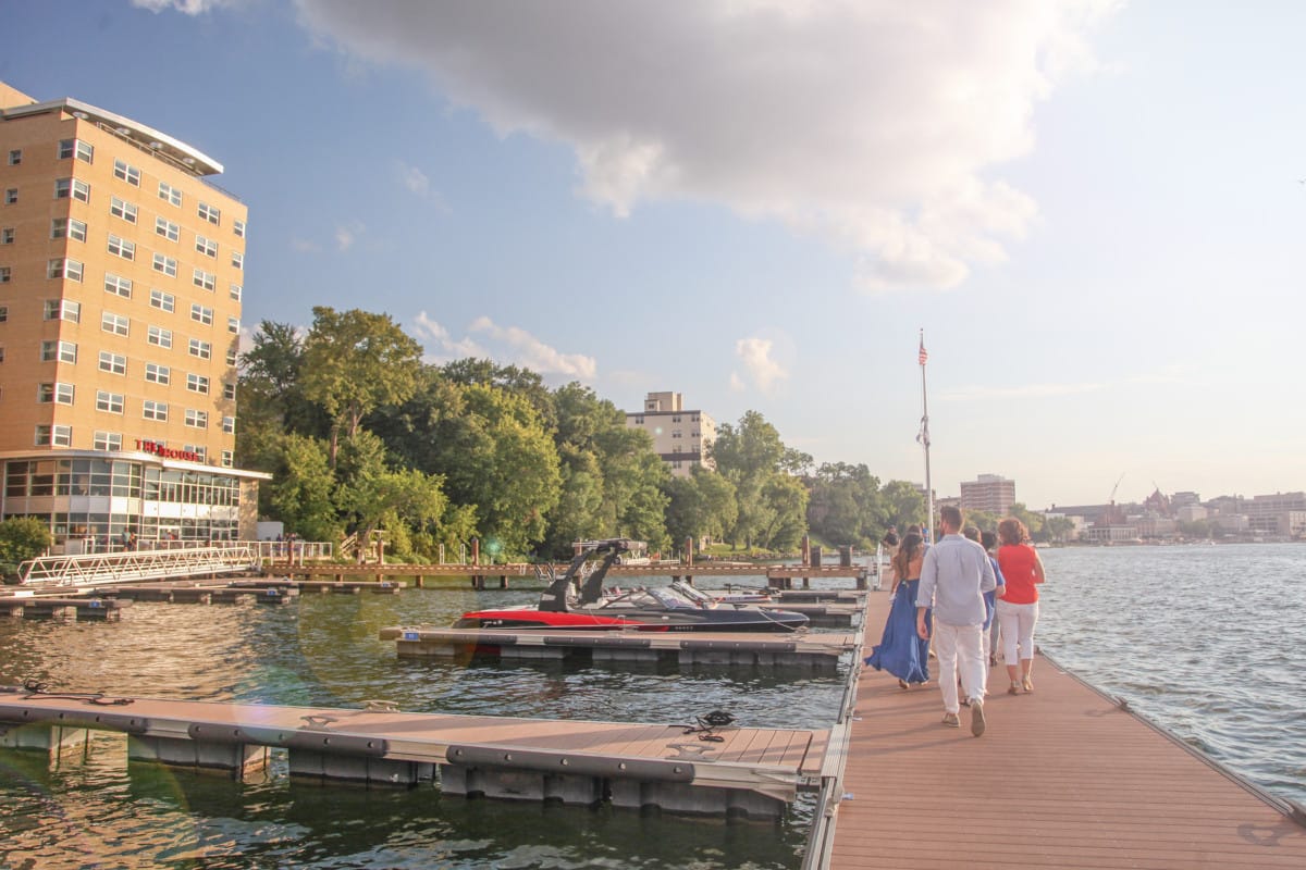 A group walks along a wooden dock lined with boats, with a hotel and trees in the background under a partly cloudy sky.