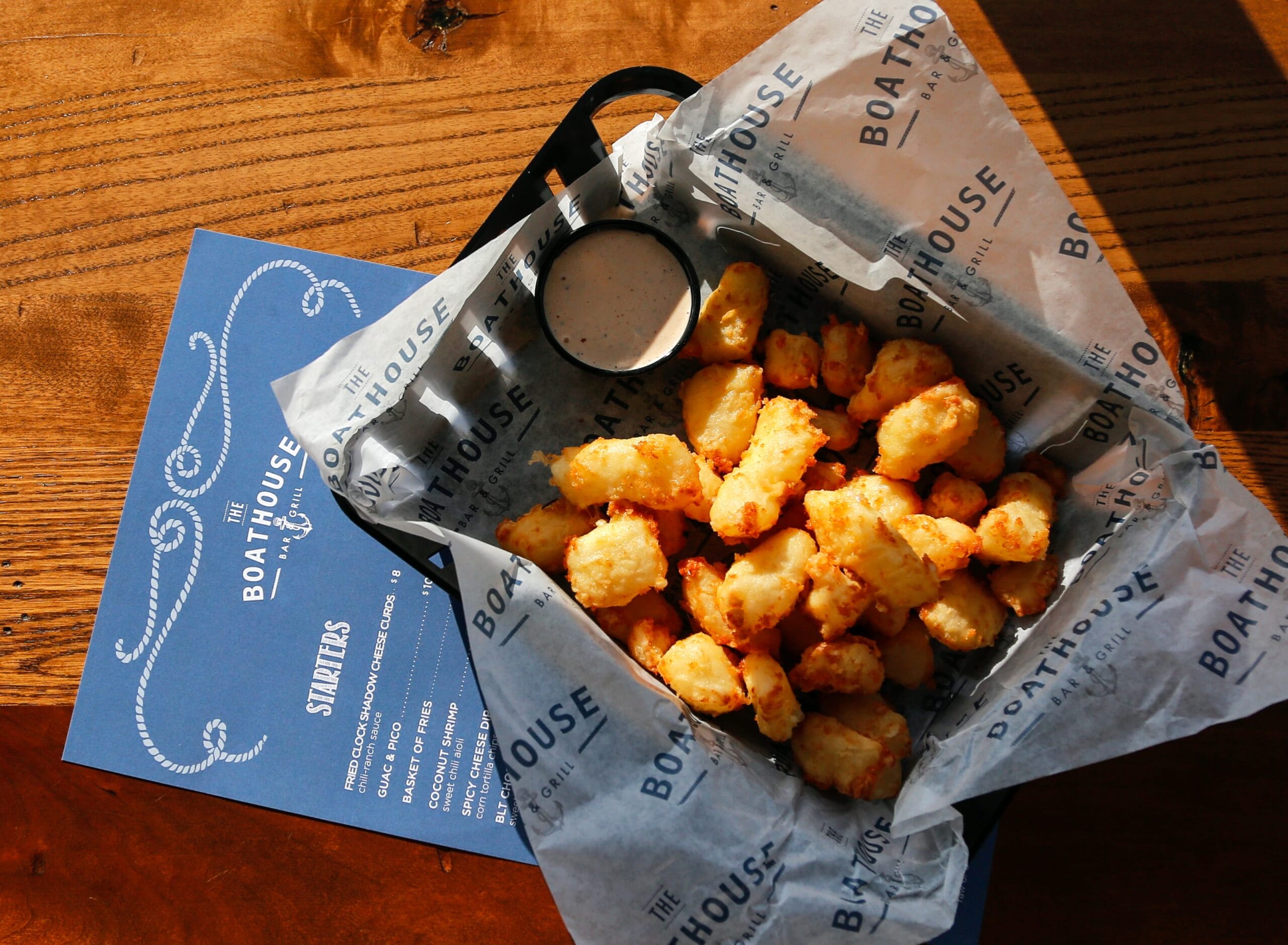 Crispy cheese curds in a basket with a side of dipping sauce, resting on a wooden table next to a menu.